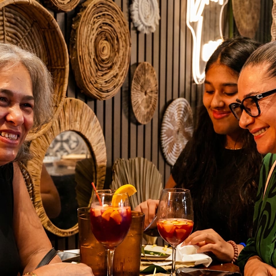 Three women smile together over colorful cocktails at a warmly lit restaurant table with woven wall decor.