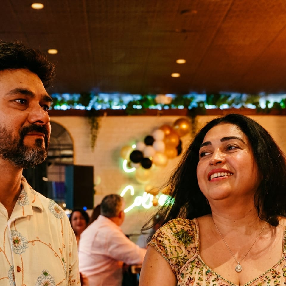 Smiling couple dressed festively at a warmly lit restaurant celebration with guests and colorful decorations behind them.