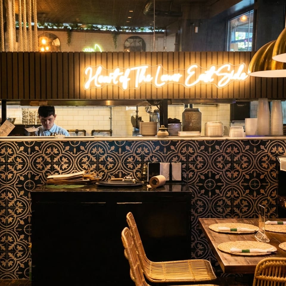 Warmly lit restaurant interior with patterned tile counter, glowing neon sign, hanging brass lamps, and set dining tables.