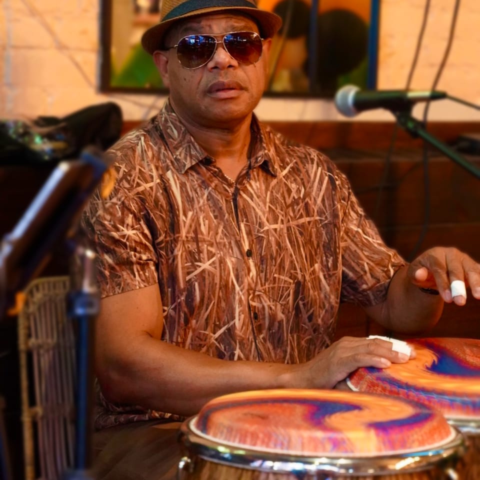 Musician wearing a fedora and sunglasses plays colorful hand drums in a warmly lit restaurant setting.