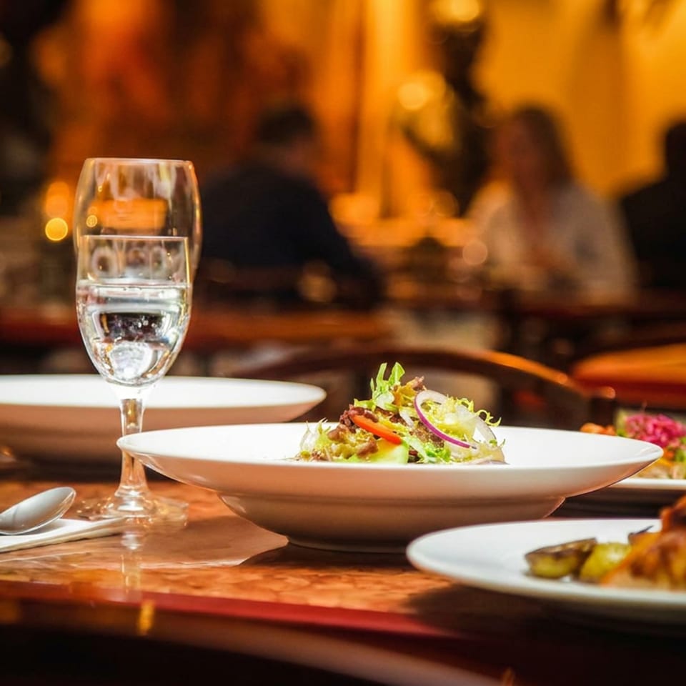 Elegant restaurant table set with plated dishes, wine and water glasses, and warmly lit diners in the background.