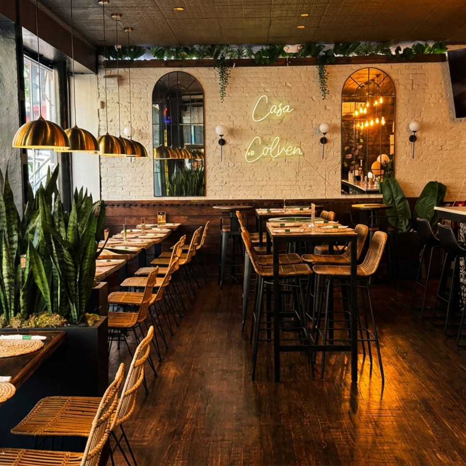 Warm, plant-filled restaurant interior with pendant lights, wooden tables, patterned bar seating, and Casa Calavera signage.