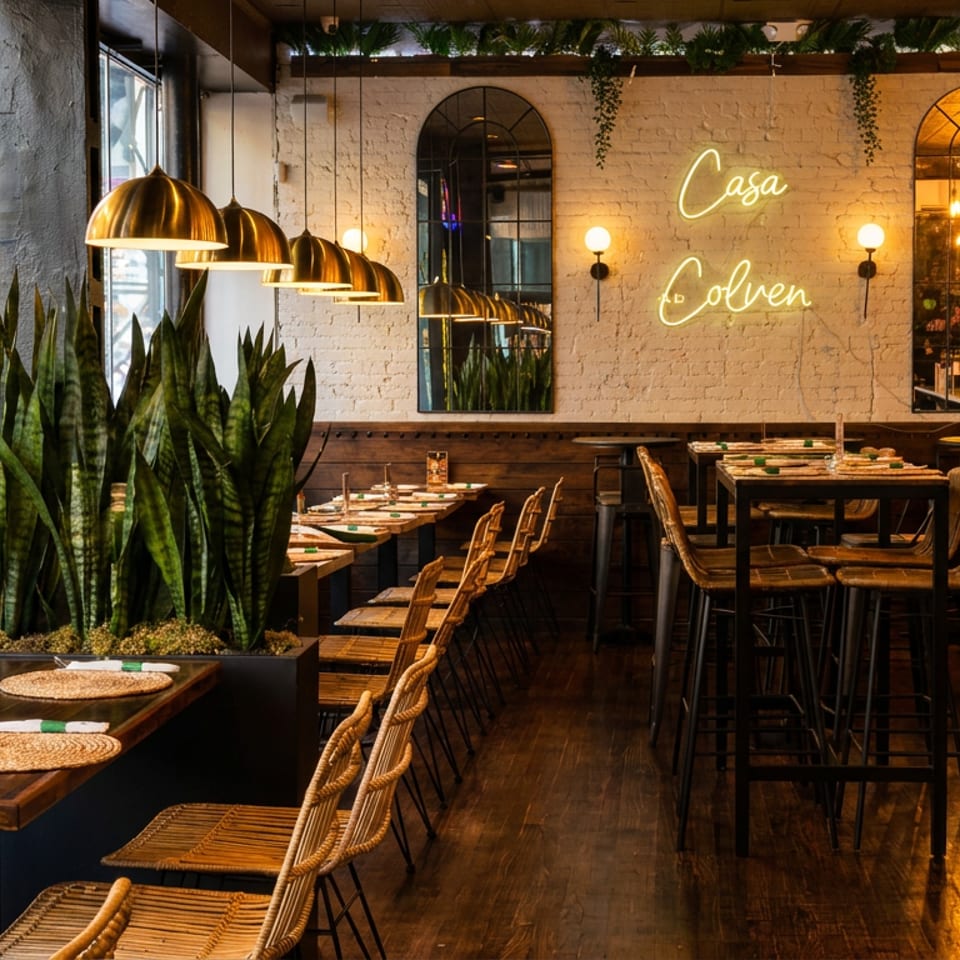 Warmly lit Casa Colven restaurant interior with bamboo seating, brass pendant lights, plants, set tables, and a neon wall sign.
